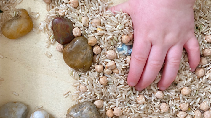 A toddler's hand scooping up rice in a wood sensory bin.